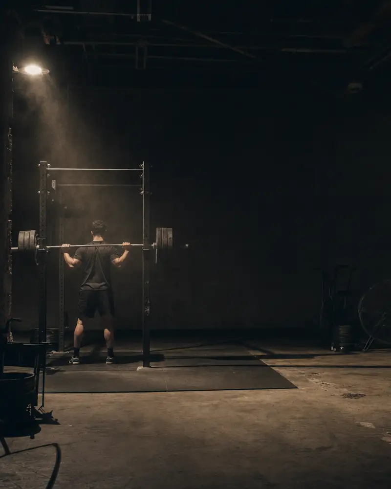 A lifter stands in a power rack with a loaded barbell on his back, about to unrack a squat in a dim gym.